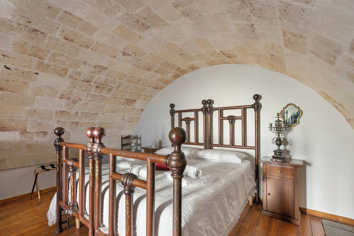 A bedroom with a wooden four-poster bed, a nightstand with a mirror, and a brick ceiling.