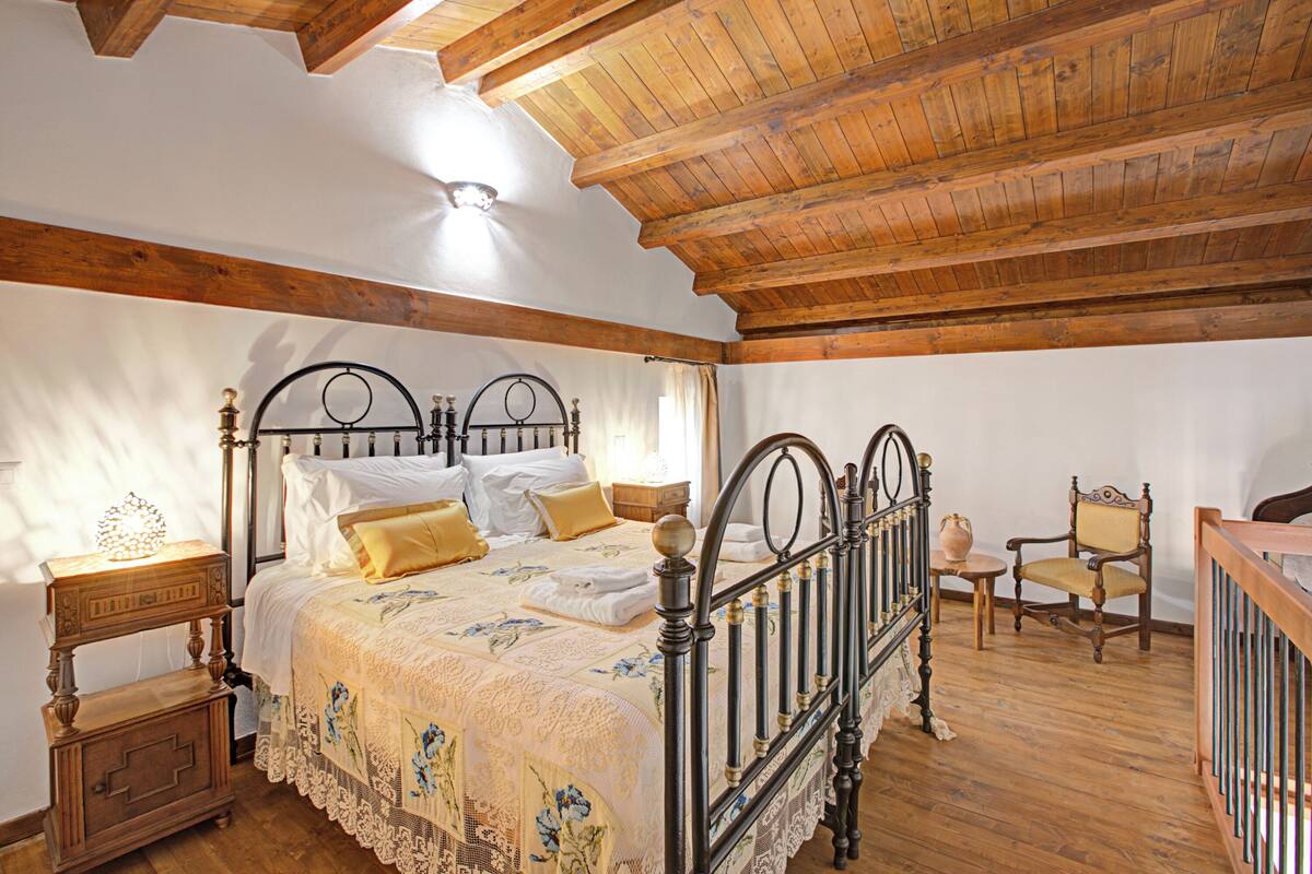 A bedroom with a wooden ceiling, a four-poster bed, a nightstand, a chair, and a small table.