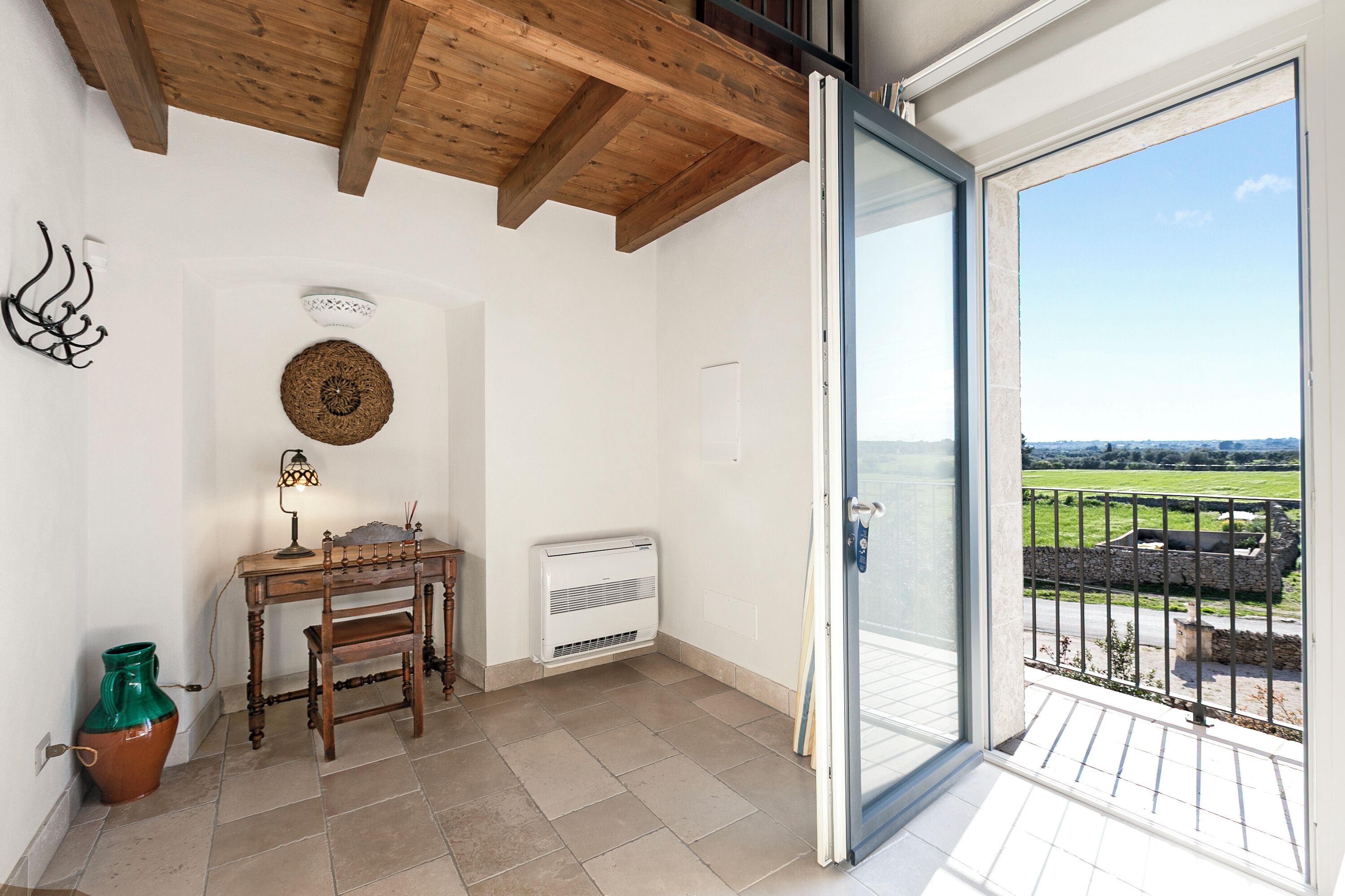 Una habitación con techo de madera, una mesa con lámpara, un radiador y vistas a un campo verde a través de un ventanal.