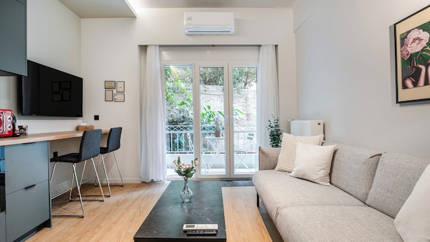 Ein modernes Wohnzimmer mit einer Couch, einem Couchtisch und einem Fernseher. Es gibt eine Küchenzeile mit einer Arbeitsplatte und Stühlen. Eine Glastür öffnet sich zu einem Balkon mit Pflanzen.