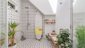 Bany compacte amb un paviment de rajoles de disseny geomètric, una dutxa amb una taula de surf i diverses plantes en testos.