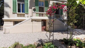 A two-story building with green shutters, a stone staircase, and a garden with a flowering plant.