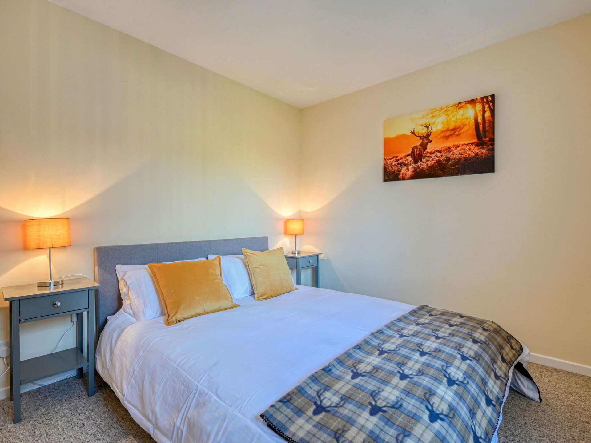 A neatly made bed with white linens and two gold pillows, flanked by two bedside tables with lamps, in a room with a single wall-mounted painting.