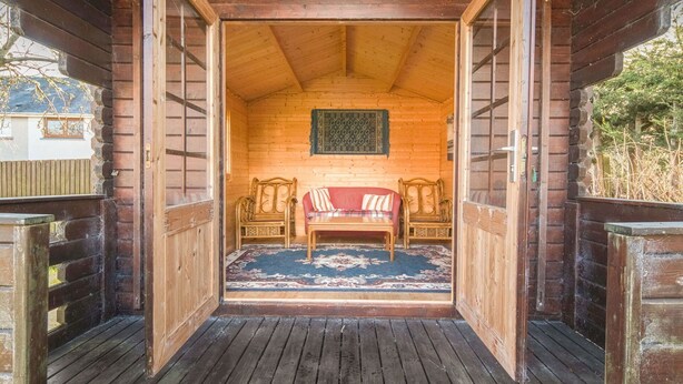 Interior de una cabaña de madera con un sofá rojo, dos sillas de madera, un tapete estampado y un cuadro en la pared.