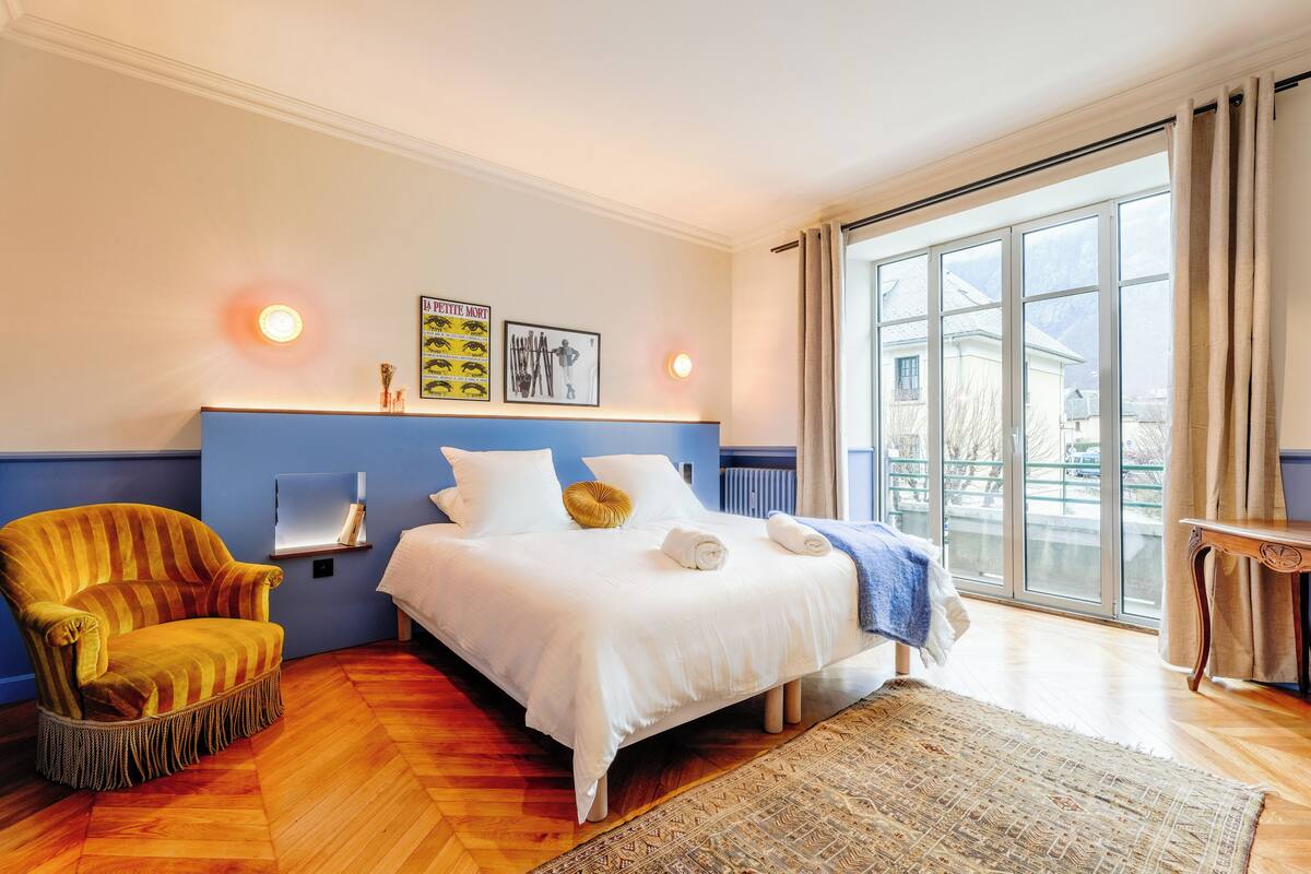 A bedroom with a bed, a chair, a window, and a wall-mounted shelf.