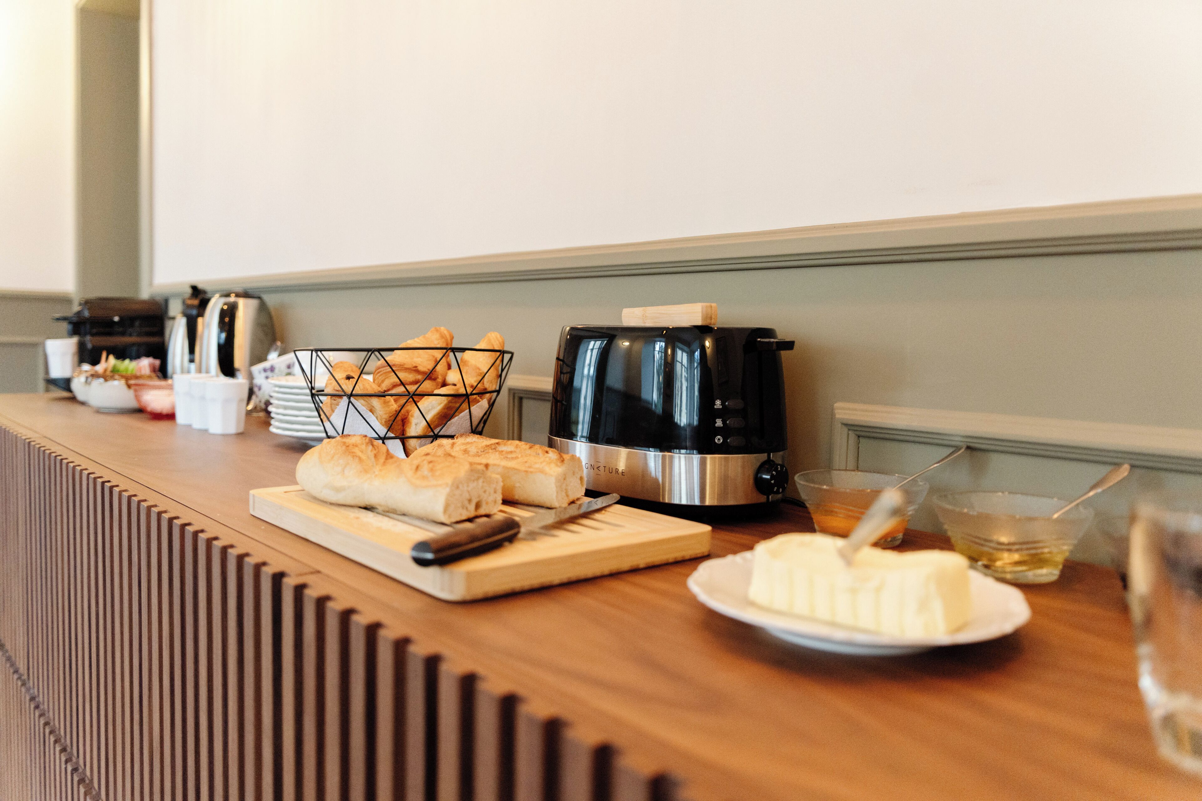 A kitchen counter with a bread basket, toaster, cutting board, knife, plate of butter, and a glass of water.