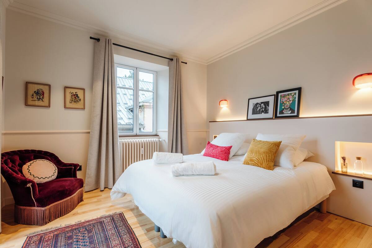 A bedroom with a large bed, a red chair, and a window with curtains.