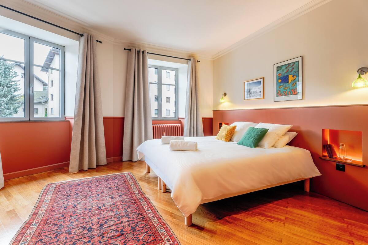 A hotel room with a large bed, a red accent wall, a window with curtains, and a small shelf with decorative items.
