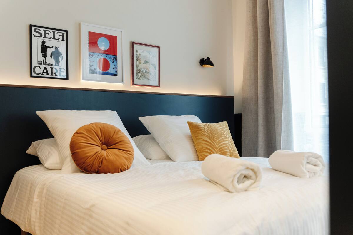 A neatly made bed with white and brown pillows, a framed picture of a person, and a wall-mounted light fixture.