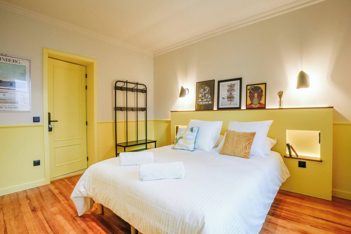 A hotel room with a bed, a bedside table, a wall-mounted light, and a door.
