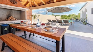 Eine Terrasse im Freien mit Holztisch, der zum Essen gedeckt ist, einem Grill und Lounge-Sesseln unter einem Sonnenschirm.