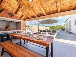 An outdoor patio with a wooden table set for dining, a barbecue grill, and lounge chairs under an umbrella.