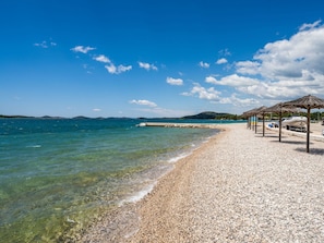 A pebble beach with clear water, a pier, and a row of thatched umbrellas.