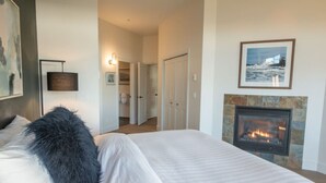 A bedroom with a bed, a black furry pillow, a fireplace, and a hallway leading to a bathroom. - Big Beach Escape – Oceanview•Walk to Trail•Fireplace (Ucluelet)