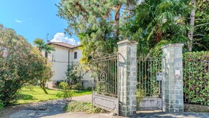 Une entrée fermée par un portail, avec des piliers en pierre et un portail en métal, qui mène à un bâtiment doté d’un toit en tuiles et entouré de verdure.