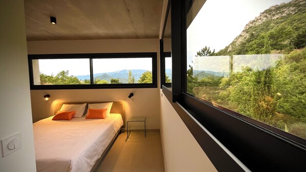 Ein Schlafzimmer mit einem Bett, einem Nachttisch, einem Fenster und Blick auf eine Berglandschaft.