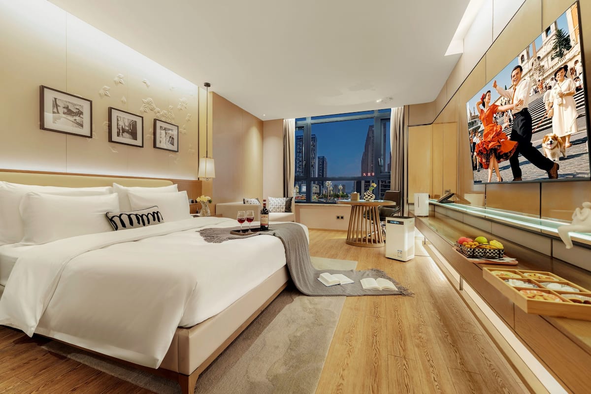 A modern hotel room with a large bed, a dining area with a view, and a cityscape visible through the window.