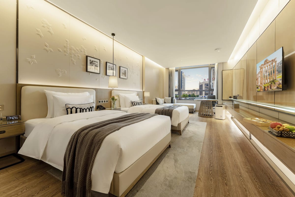 A modern hotel room with two beds, a large window with a city view, a flat-screen TV, and a breakfast bar with fruit.