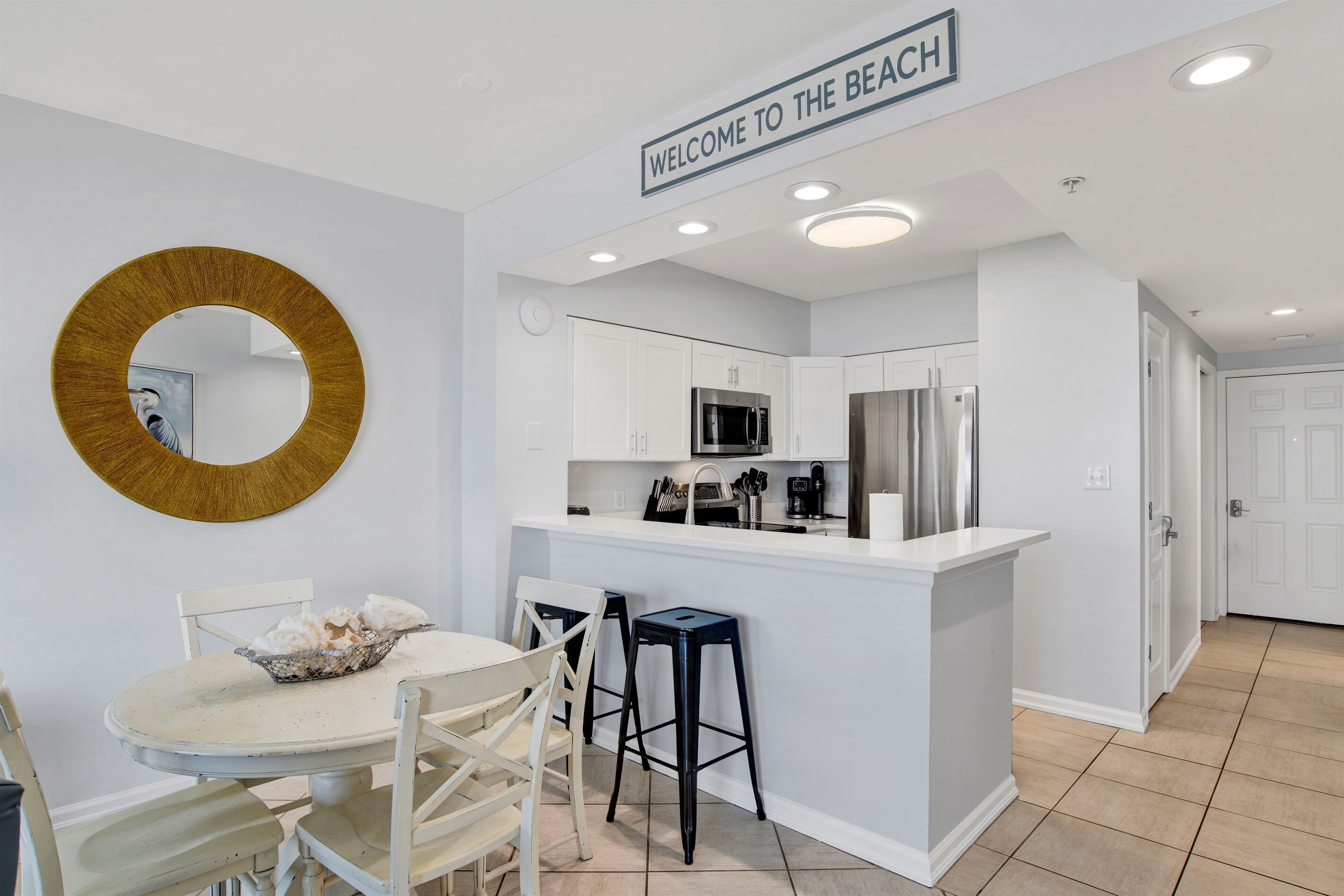 A modern kitchen with white cabinetry, a central island, and a "Welcome to the Beach" sign.