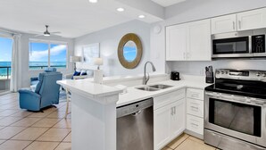 Una cocina moderna con armarios blancos, lavavajillas integrado, estufa y microondas. Hay un ventanal con vista al océano.