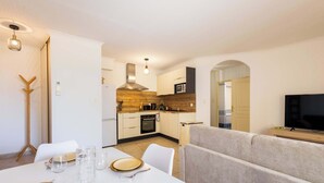 A modern kitchen with white cabinets, a microwave, and a stove. There is a dining area with a white table set for four, and a grey sofa in the background. - Valras’Loc – Rated apartment with pool & quiet terrace (Valras-Plage)