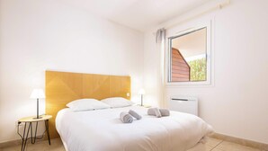 A neatly made bed with white linens, a wooden headboard, a bedside table with a lamp, and a window with a view of greenery. - Valras’Loc – Rated apartment with pool & quiet terrace (Valras-Plage)
