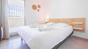 Una cama bien hecha con toallas, cabecero de madera, mesita de noche y vistas a través de una ventana con persianas.
