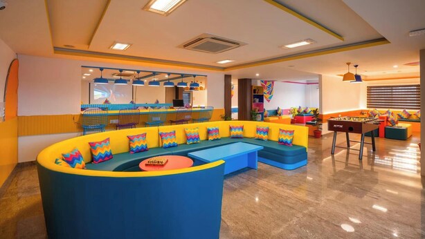 Una hall vivace con un divano blu curvo, un tavolo da calcio e un'area bar con sedie colorate e un bancone.