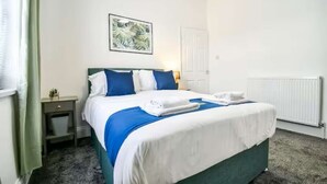 A neatly made bed with white and blue bedding, a small bedside table with a lamp, a framed picture on the wall, and a radiator. - Comfortable house in Hartlepool (Hartlepool)