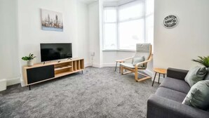 A modern living room with a flat-screen TV on a wooden stand, a grey sofa, a corner window, and a small table with a chair. - Comfortable house in Hartlepool (Hartlepool)