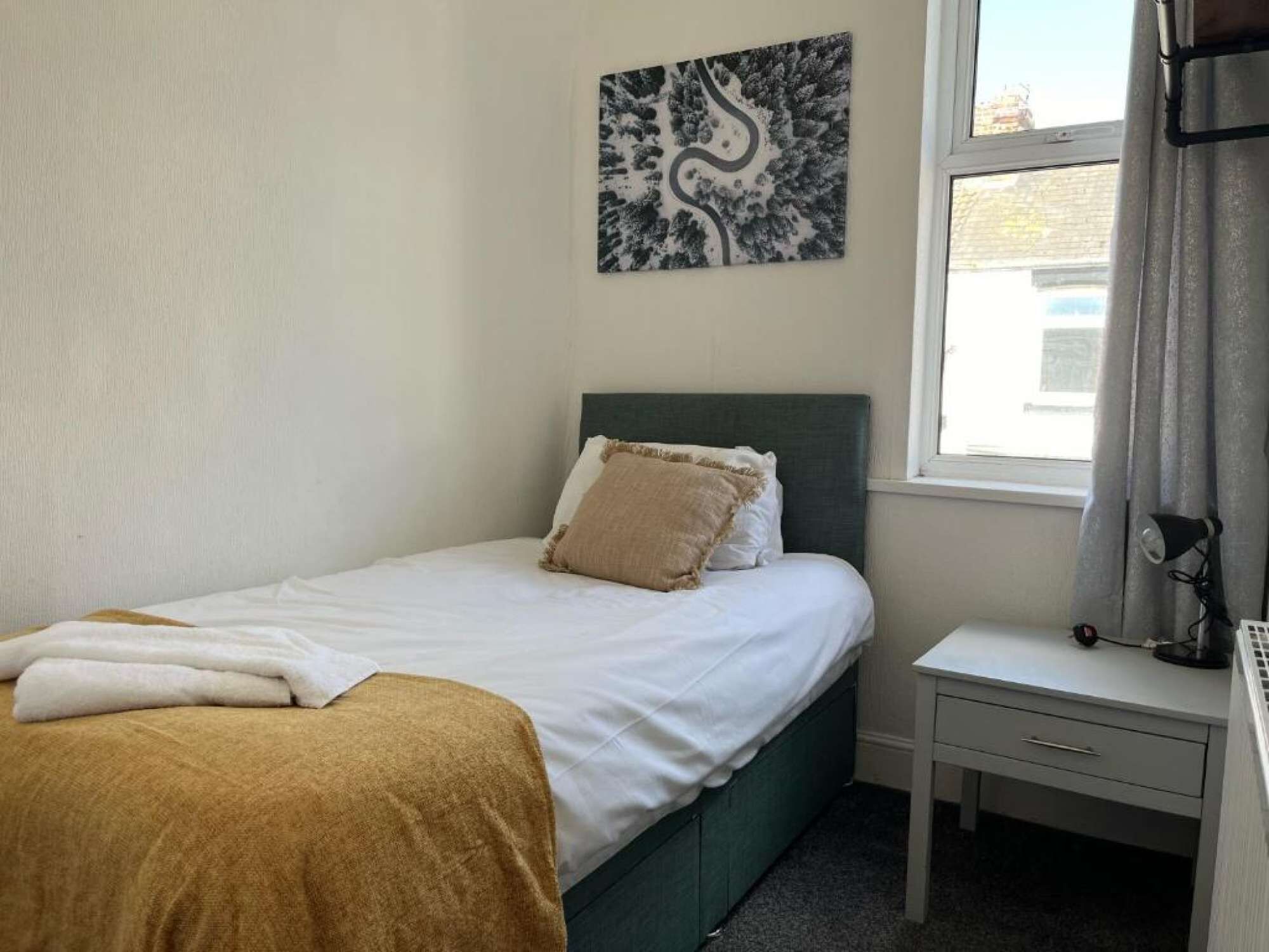 Un dormitorio con cama, mesita de noche, lámpara y ventana con cortinas.