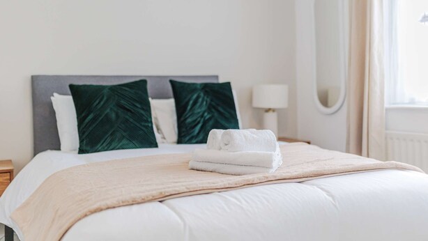 Een keurig opgemaakt bed met een beige deken en twee kussens, een bedlampje en een spiegel aan de muur.