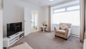 Een moderne woonkamer met een flatscreen televisie, een beige fauteuil en een groot raam met gordijnen.