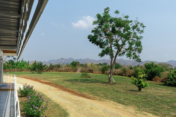 A view from a building's porch overlooking a dirt path leading to a grassy area with a tree and distant mountains. - Narucha Farmstay (Kanchanaburi)