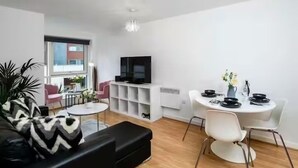 Un salon moderne avec un canapé noir, une petite table ronde, une table à manger blanche avec des chaises, un téléviseur posé sur un meuble blanc et une grande fenêtre.