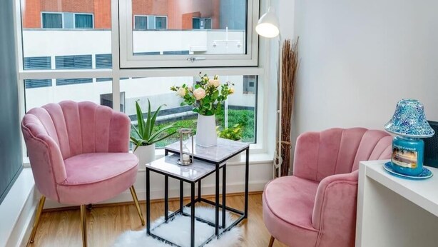 Une pièce avec deux chaises roses, un vase blanc rempli de fleurs, une petite table et une lampe bleue posée sur une étagère blanche.