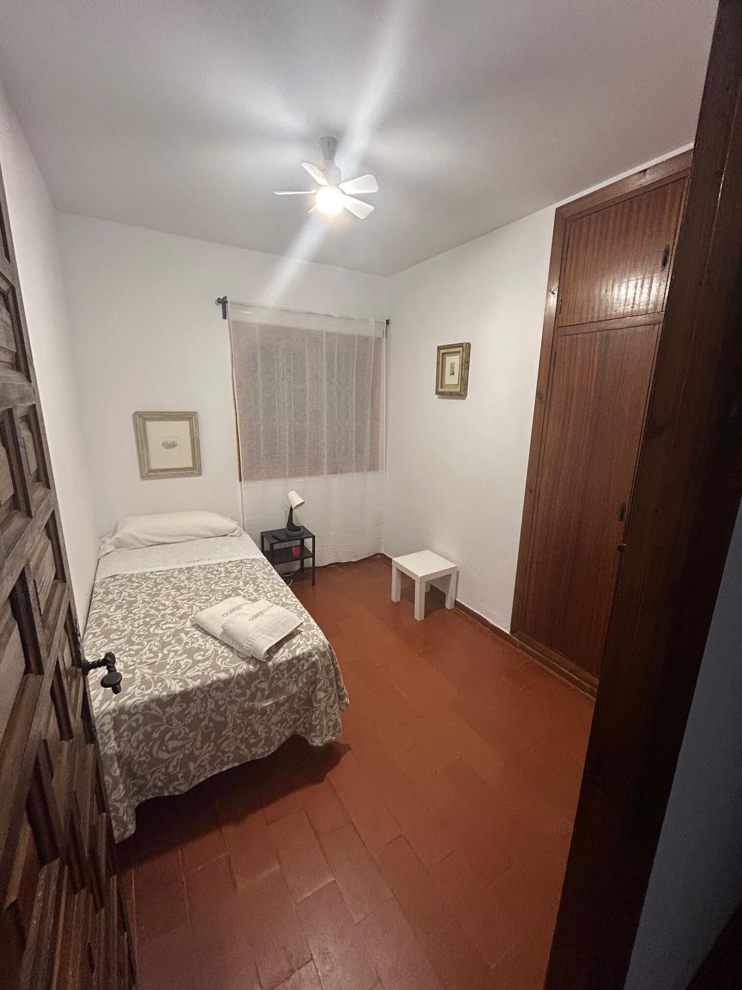 Ein Schlafzimmer mit einem Bett, einem kleinen Nachttisch, einem Hängeventilator, einem Fenster mit Vorhängen und einem Holzschrank.