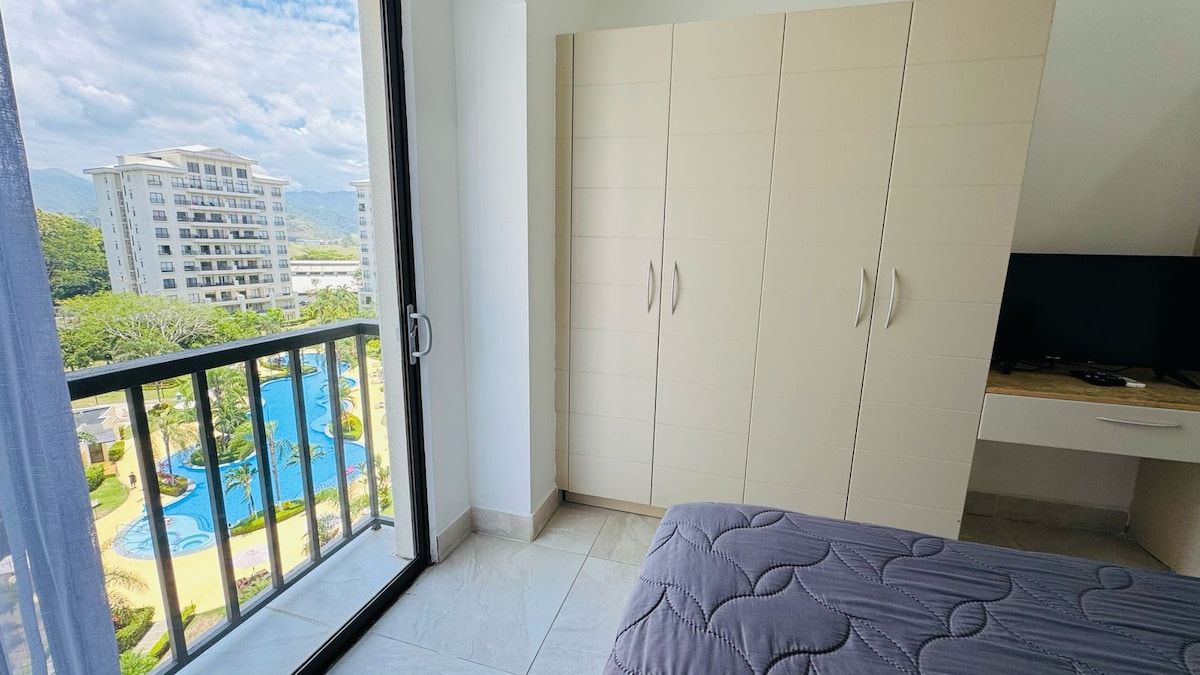 Ein Schlafzimmer mit Bett, Kleiderschrank und Fernseher. Es gibt einen Balkon mit Blick auf einen Swimmingpool und Gebäude.
