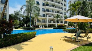 A hotel pool area with lounge chairs and umbrellas, surrounded by palm trees and a multi-story building. - Jaco Bay 5701 (Jaco Beach)