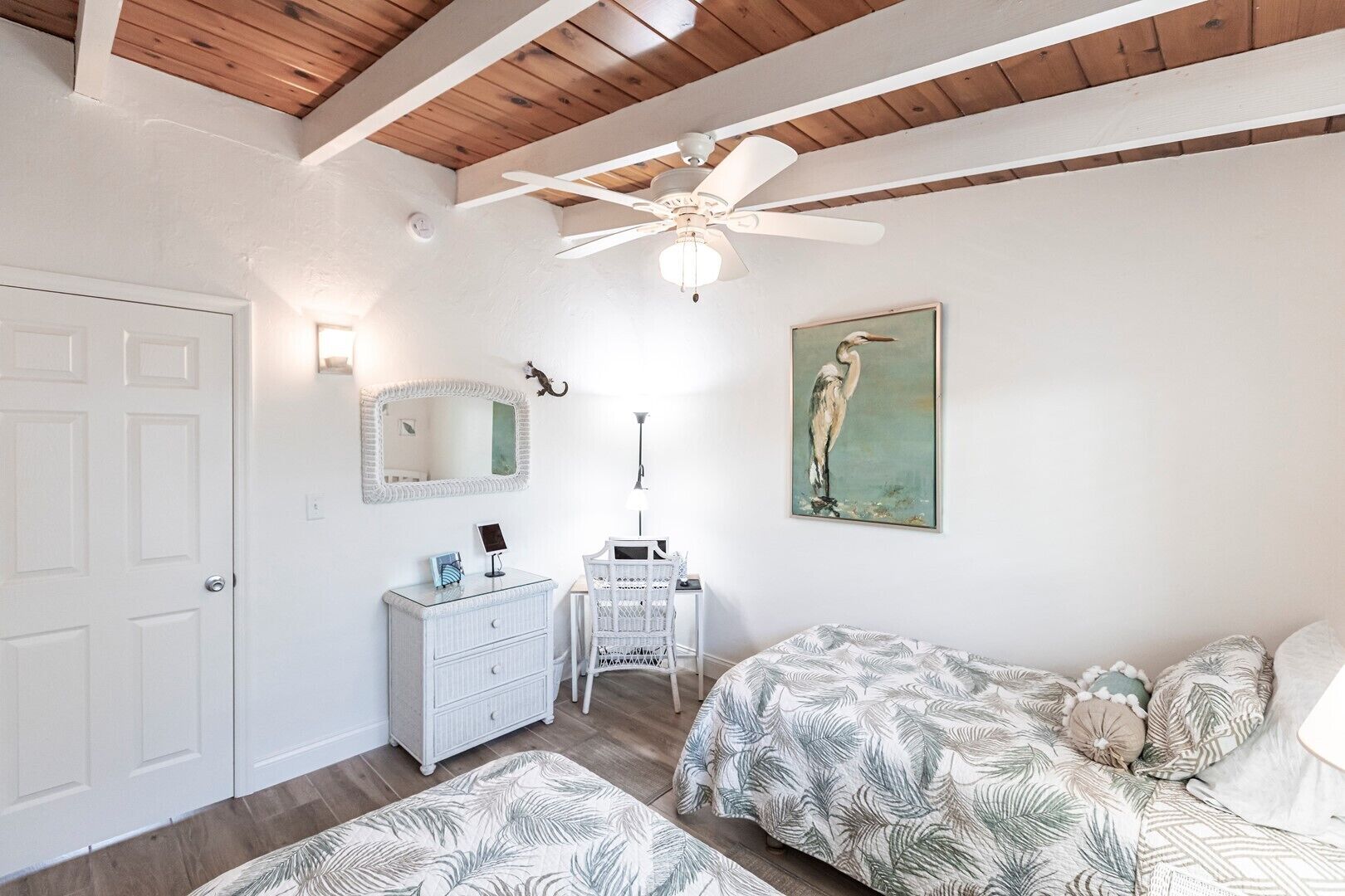 Ein Schlafzimmer mit weißem Schminktisch, einem Bett mit blattgemustertem Überwurf, einem Deckenventilator und einem Wandbild eines Vogels.