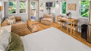 Un dormitorio con una cama grande, un sofá, una mesita y un escritorio con sillas. Hay ventanas que ofrecen vistas a zonas verdes.