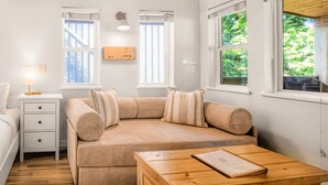 Una habitación acogedora con un sofá color beige, una mesa de madera, una mesita de noche con cajones y dos ventanas con cortinas.