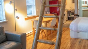 A room with a wooden floor, a grey sofa, a ladder, a dining table with chairs, and a bed.