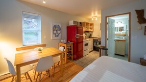 Una habitación compacta con una cama, una mesa de comedor con sillas, una kitchenette con un refrigerador rojo y una ventana con persianas.