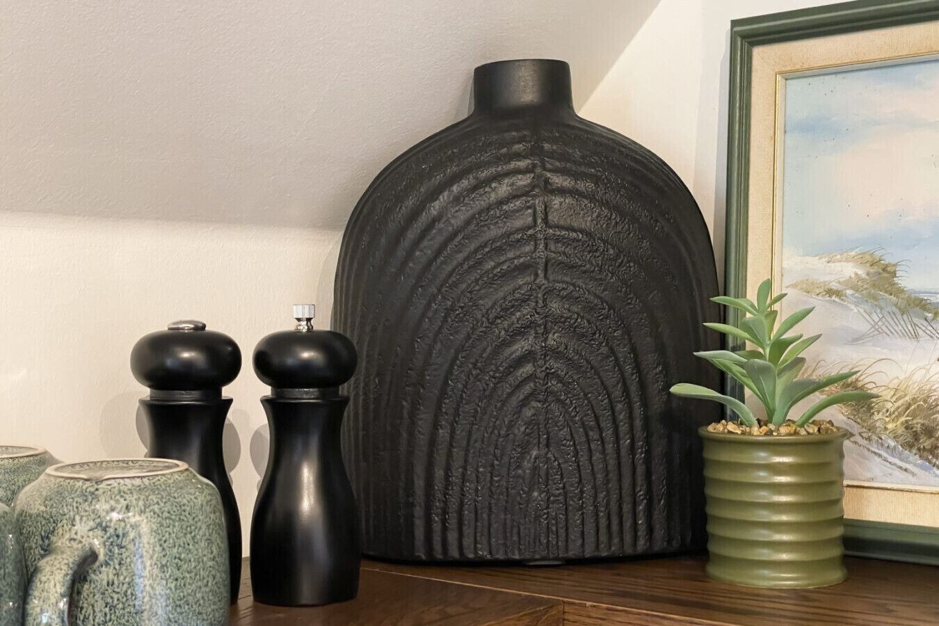 A room with a large black vase, two black pepper shakers, a green plant in a pot, and a framed painting on the wall.