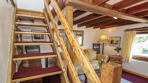 Un escalier en bois, dont les marches sont recouvertes d’une moquette rouge, mène à un salon où se trouvent un canapé, une lampe et une fenêtre ornée de rideaux.