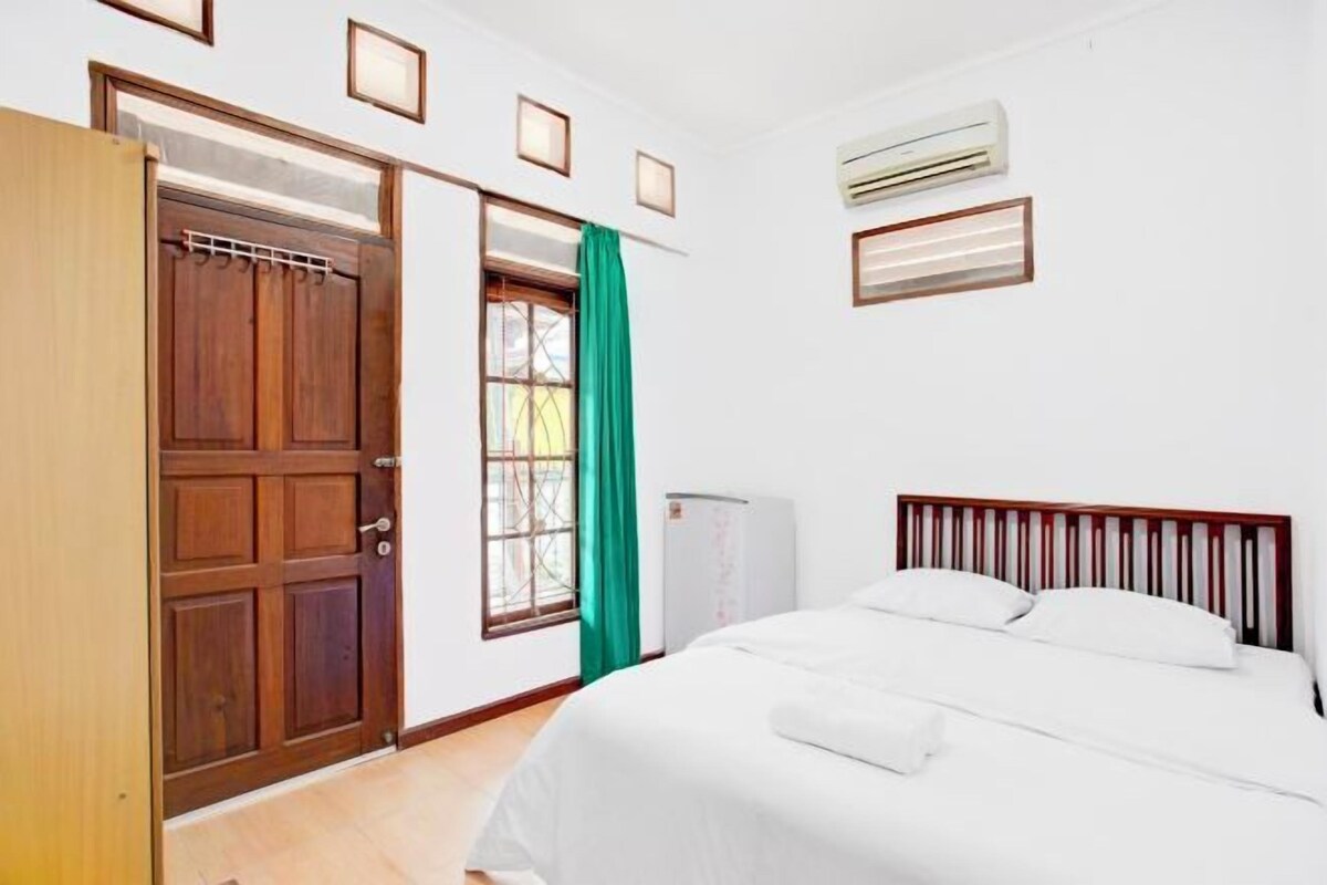 A bedroom with a wooden wardrobe, a bed with white bedding, a window with green curtains, and an air conditioning unit on the wall.