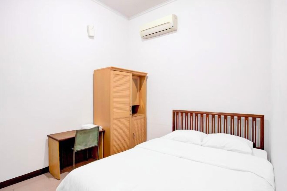 A bedroom with a bed, a wooden wardrobe, a desk with a chair, and an air conditioning unit.