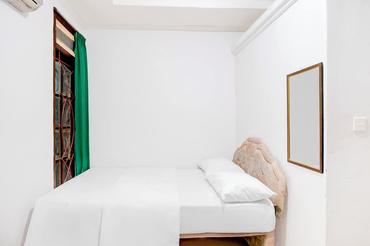 A small, white-walled bedroom with a single bed, a green curtain, and a framed mirror on the wall.