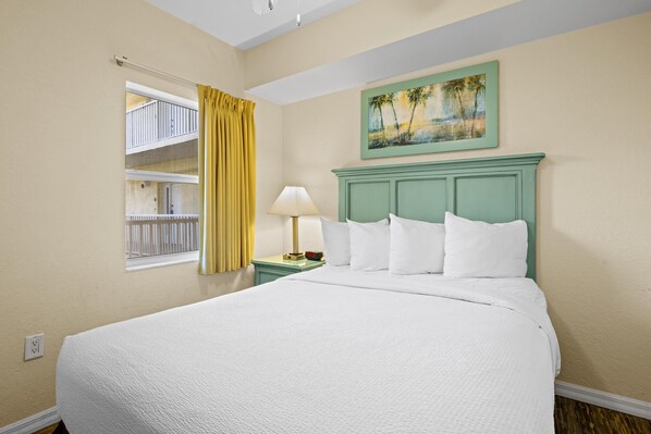 Habitación de hotel con una cama grande, una mesita de noche con lámpara, un cuadro de palmeras y una ventana con cortinas amarillas.